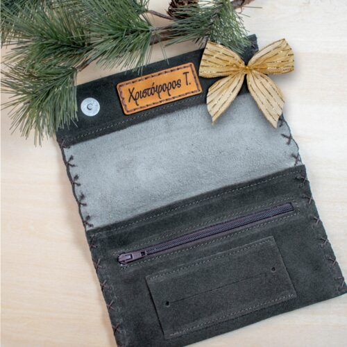 Ανοιχτή δερμάτινη θήκη καπνού σε στυλ φακέλου, φτιαγμένη από σκούρο σουέτ. Το εσωτερικό της είναι ανοιχτό γκρι και στην επάνω πλευρά υπάρχει ραμμένη δερμάτινη ετικέτα σε ανοιχτό καφέ χρώμα με χαραγμένο το όνομα «Χριστόφορος Τ.». Στο κάτω μέρος της θήκης υπάρχει μία οριζόντια τσέπη με φερμουάρ και από κάτω μια επίπεδη θήκη για χαρτάκια. Γύρω από τις άκρες φαίνεται χειροποίητο ράψιμο. Η θήκη είναι τοποθετημένη σε ανοιχτό ξύλινο φόντο, διακοσμημένη με κλαδιά πεύκου και ένα χρυσό φιόγκο στην επάνω δεξιά γωνία, δίνοντας μια εορταστική αίσθηση.