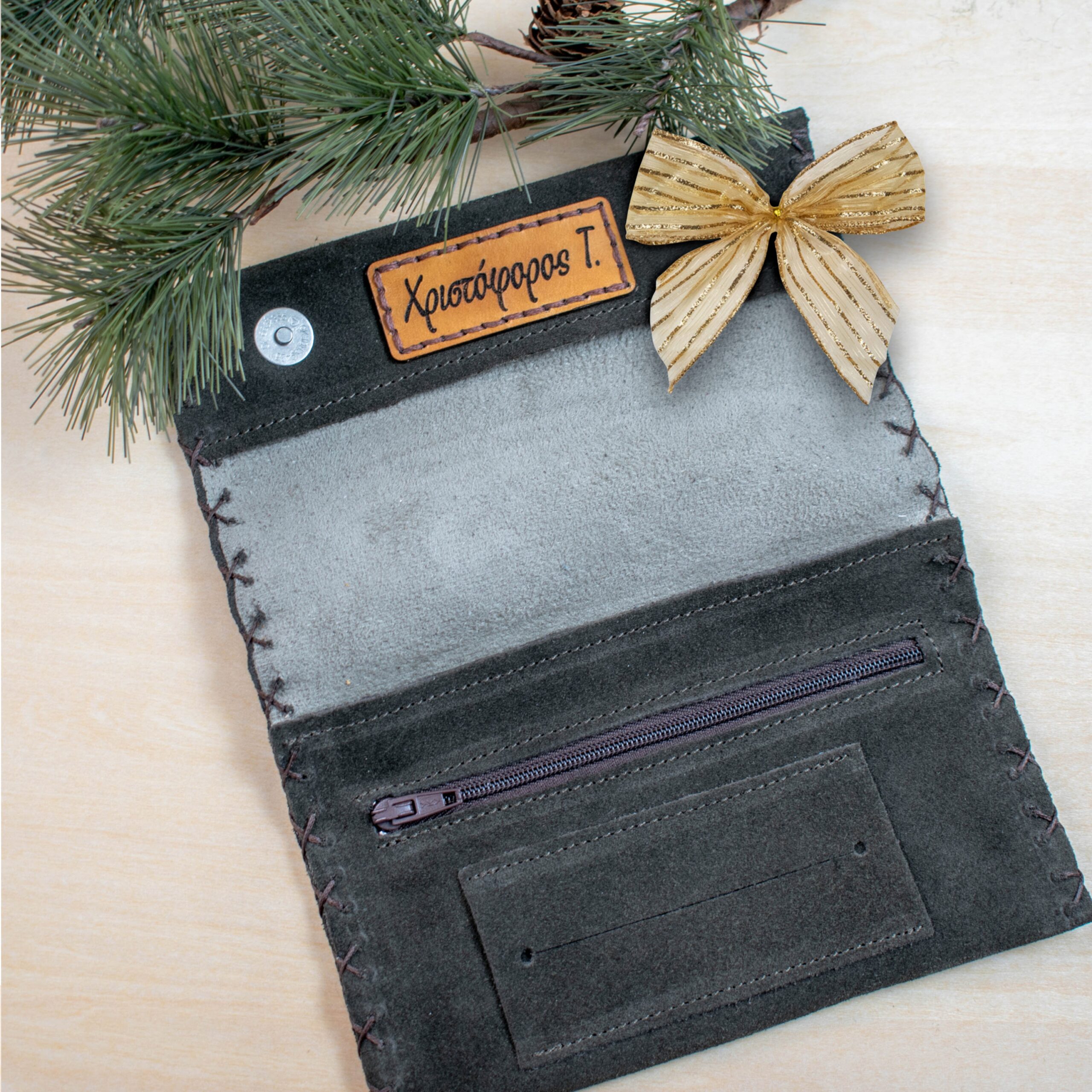 Ανοιχτή δερμάτινη θήκη καπνού σε στυλ φακέλου, φτιαγμένη από σκούρο σουέτ. Το εσωτερικό της είναι ανοιχτό γκρι και στην επάνω πλευρά υπάρχει ραμμένη δερμάτινη ετικέτα σε ανοιχτό καφέ χρώμα με χαραγμένο το όνομα «Χριστόφορος Τ.». Στο κάτω μέρος της θήκης υπάρχει μία οριζόντια τσέπη με φερμουάρ και από κάτω μια επίπεδη θήκη για χαρτάκια. Γύρω από τις άκρες φαίνεται χειροποίητο ράψιμο. Η θήκη είναι τοποθετημένη σε ανοιχτό ξύλινο φόντο, διακοσμημένη με κλαδιά πεύκου και ένα χρυσό φιόγκο στην επάνω δεξιά γωνία, δίνοντας μια εορταστική αίσθηση.