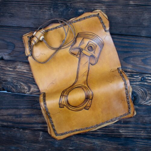 Handcrafted Leather Tobacco Case with Piston Pyrography This is a unique, handcrafted leather tobacco case with a strong automotive, mechanical, or engine-inspired aesthetic. It is perfect for someone who appreciates speed, engineering, or a rugged, industrial style. Materials and Texture Material: The case is made from genuine, substantial leather, dyed a warm, natural tan or amber color. Feel: The leather is smooth, thick, and durable, designed to feel sturdy yet pliable. You can notice the organic, slightly uneven edges of the leather, which contribute to its rustic, handmade look. Stitching: The entire perimeter of the case is bound with a thick, braided leather cord (a whipstitch). This stitching creates a distinctive, raised, and highly tactile border all around the pouch. Design and Pyrography (The Tactile Art) The central and most prominent feature is a large design created by pyrography (the art of burning/etching a design into the leather). This burning process leaves a smooth, shallow indentation on the leather's surface, making the artwork fully noticeable by touch. The Piston and Connecting Rod: The etching depicts a detailed image of an engine piston and its connecting rod—essential components of an internal combustion engine. You can trace the smooth, cylindrical head of the piston at the top. The long, narrow connecting rod extends downward from the piston head, ending in a large, smooth circular opening (the bearing end). The lines representing the structure, joints, and piston rings are all etched, allowing the user to feel the mechanical details and the overall shape of the engine part. Closure and Function Shape and Size: The pouch is roughly rectangular but features softly curved or irregular edges. Closure: The top corner features a simple, effective closure. A thin leather drawstring is attached to the case and wraps around a small, smooth, oval-shaped wooden bead or toggle to keep the contents secure. This mechanism is easy to locate and operate by feel. Overall Impression This piece has a gritty, cool, and technical personality. It smells subtly of natural, tanned leather and is ideal for an enthusiast of hot rods, motorcycles, or mechanical craftsmanship. The tactile etching turns an engine part into a unique piece of wearable art.