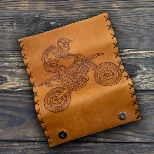 Δερμάτινη θήκη σε φυσική καφέ απόχρωση, με τετράγωνο περίπου σχήμα και ελαφρώς ακανόνιστες, χειροποίητες άκρες. Η επιφάνειά της είναι λεία, με ορατή την υφή του δέρματος. Στο κέντρο υπάρχει χαραγμένο σχέδιο που απεικονίζει έναν αναβάτη motocross με κράνος, τη στιγμή που οδηγεί μια off-road μοτοσυκλέτα σε κίνηση, με τους τροχούς και τις λεπτομέρειες της μηχανής καθαρά αποτυπωμένες. Περιμετρικά, το δέρμα είναι ραμμένο στο χέρι με σκούρα κορδόνια που περνούν μέσα από τρύπες, δημιουργώντας εμφανείς σταυρωτές ραφές. Στο κάτω μέρος υπάρχουν δύο μεταλλικά κουμπώματα που κλείνουν τη θήκη. Το αντικείμενο αποπνέει χειροποίητο, rustic χαρακτήρα και αίσθηση ανθεκτικότητας.