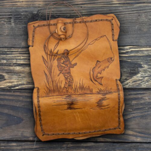 Χειροποίητη δερμάτινη θήκη σε ζεστή καφέ απόχρωση, με ακανόνιστο ορθογώνιο σχήμα και εμφανή φυσική υφή δέρματος. Περιμετρικά είναι ραμμένη στο χέρι με χοντρό δερμάτινο κορδόνι, δημιουργώντας έντονες ραφές που τονίζουν τον rustic χαρακτήρα της. Στο επάνω μέρος διαθέτει δερμάτινο κορδόνι δεσίματος με ξύλινο κουμπί για κλείσιμο. Στο κέντρο της επιφάνειας υπάρχει χαραγμένο σχέδιο που απεικονίζει έναν ψαρά να ρίχνει το καλάμι του σε λίμνη ή ποτάμι, με το ψάρι να αναδύεται από το νερό. Στο φόντο διακρίνονται καλάμια και αντανακλάσεις στο νερό. Το αντικείμενο αποπνέει φυσικότητα, χειροποίητη ποιότητα και σύνδεση με τη φύση και το ψάρεμα.