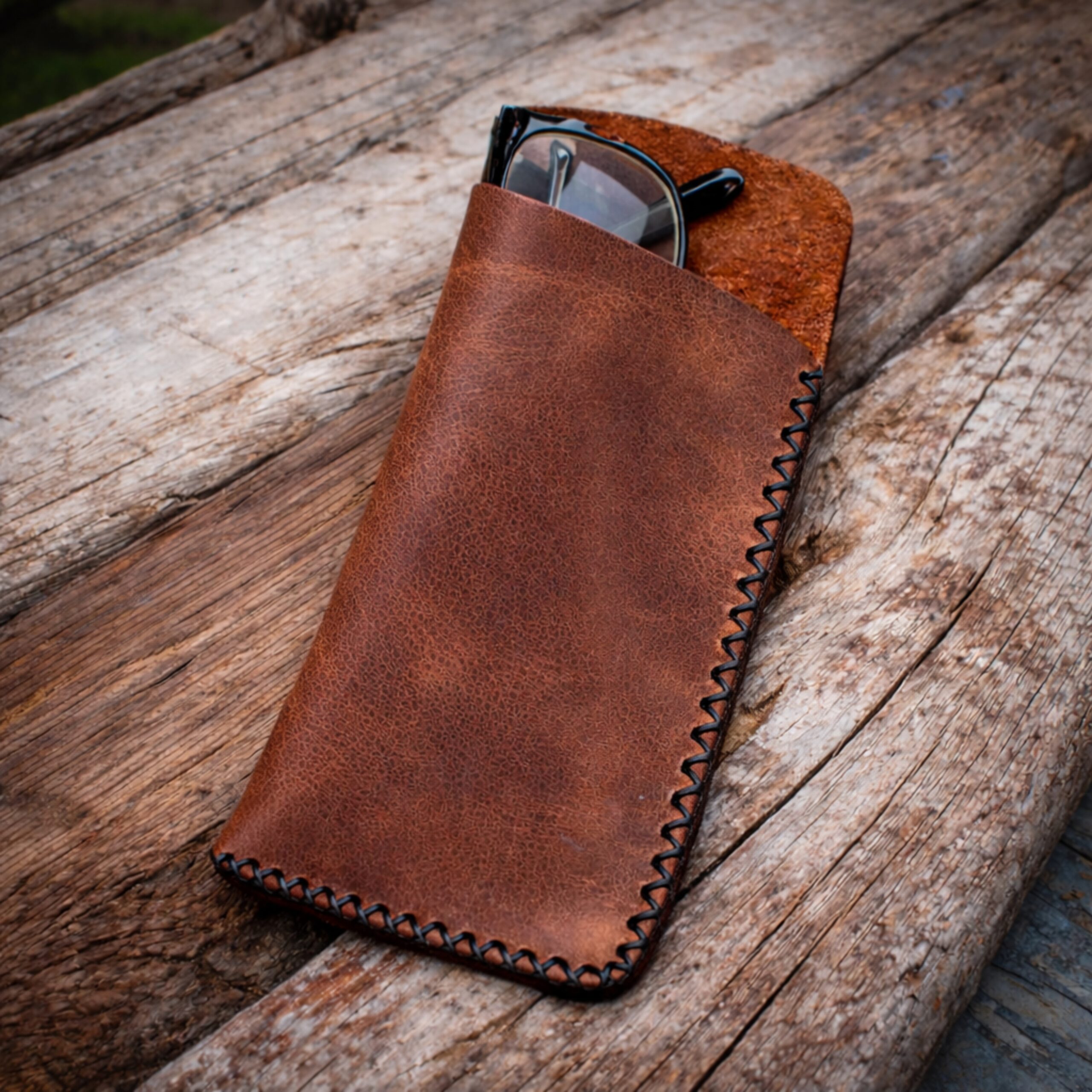 Χειροποίητη δερμάτινη θήκη γυαλιών σε ταμπά χρώμα με εμφανή μαύρη ραφή στο χέρι, τοποθετημένη πάνω σε φυσικό ξύλο