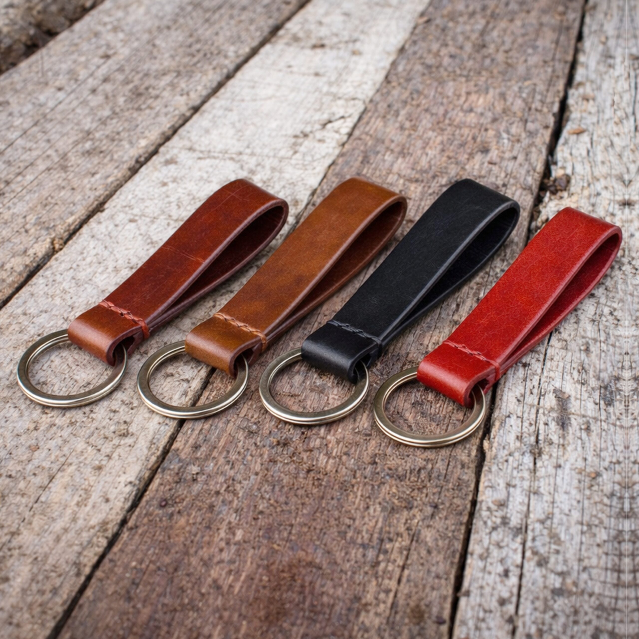 Τέσσερα χειροποίητα δερμάτινα μπρελόκ με ραφή σε χρώματα ταμπά, καφέ, μαύρο και κόκκινο, τοποθετημένα σε ξύλινη επιφάνεια.